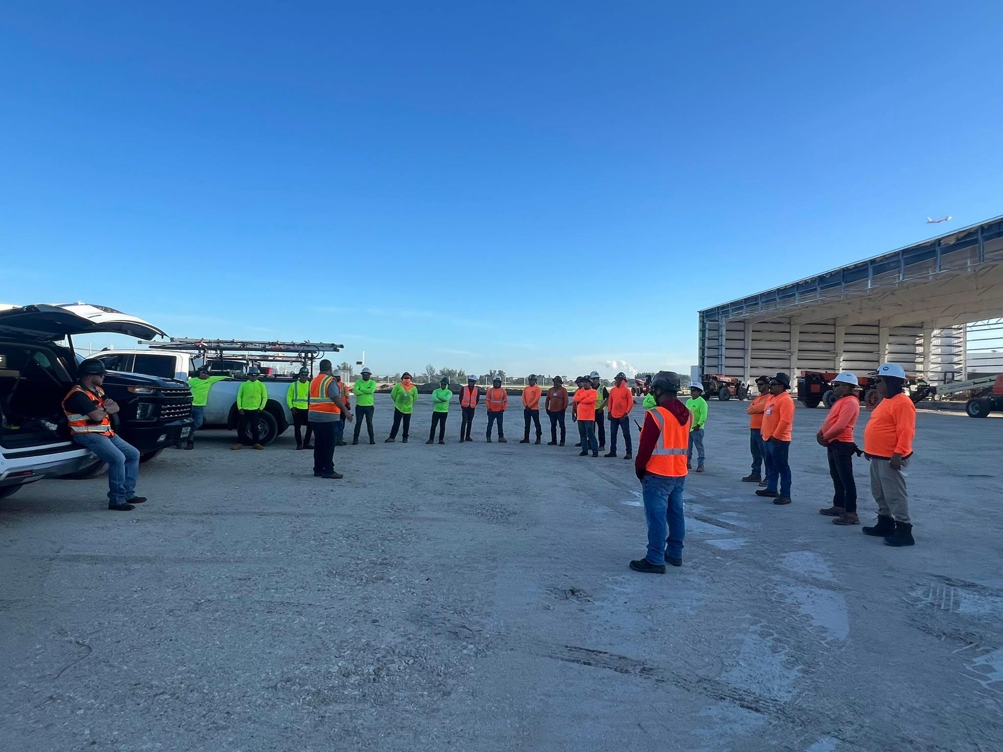Full crew in hard hats and hi-vis vests standing in a circle for a documented site safety briefing in front of a metal building