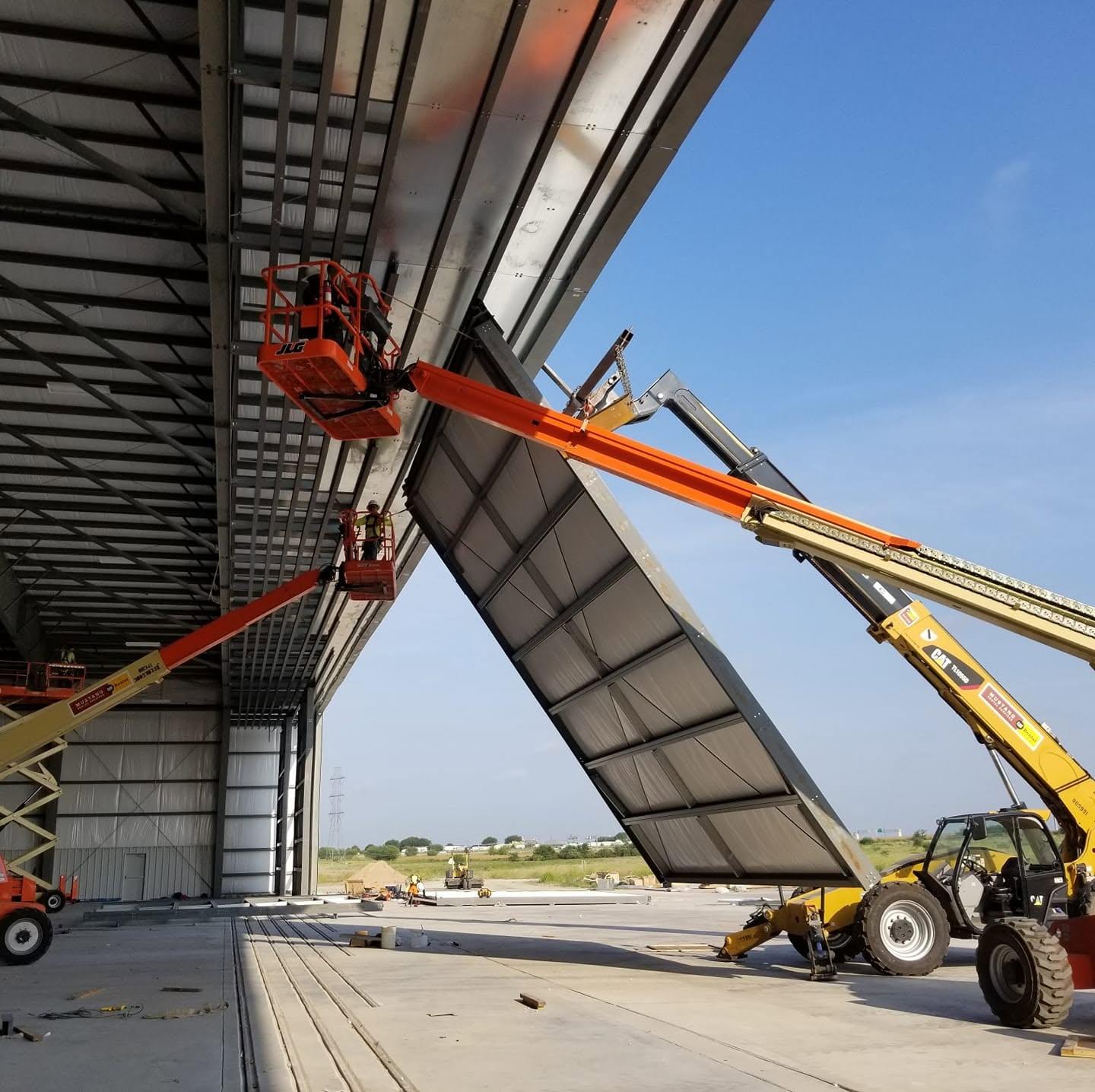 Crew member in a harness and hard hat guiding a large wall panel into place from a boom lift inside a steel building