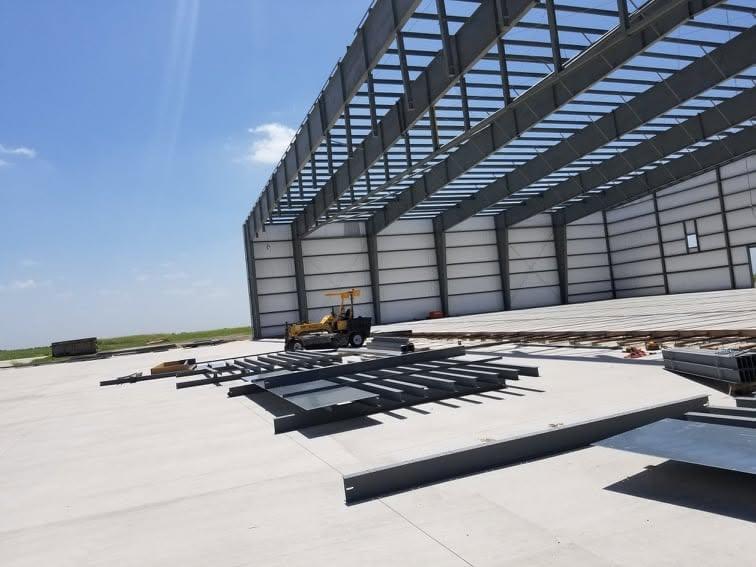 Interior of a partially erected steel building with roof purlins overhead and steel members staged across the slab