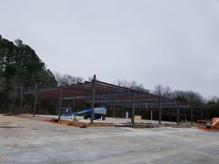 Bare steel building frame standing against a winter treeline, with columns plumbed and rafters set