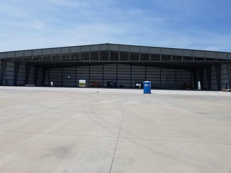 Long metal aircraft hangar with a row of bay doors, seen wide across an open concrete apron under a clear sky