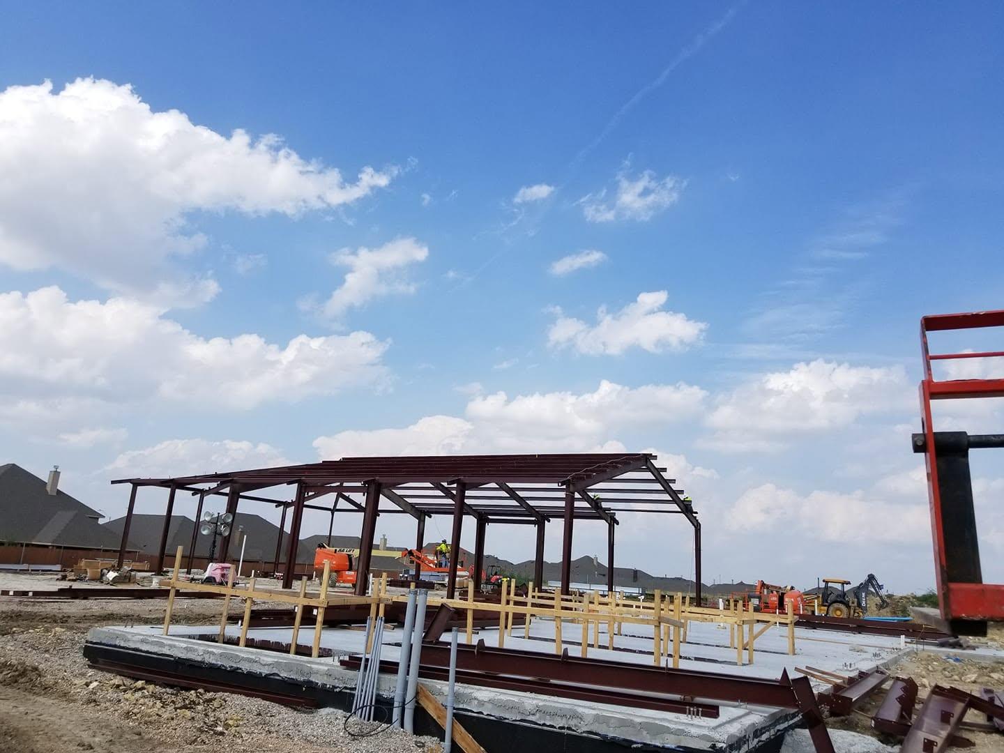 Crew erecting a large red primed steel frame, with rigid frames set and bracing going in across several bays