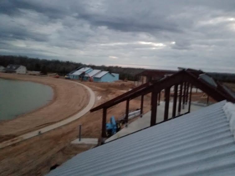Open steel pavilion structure beside a pond with metal roof panels being installed across the rafters